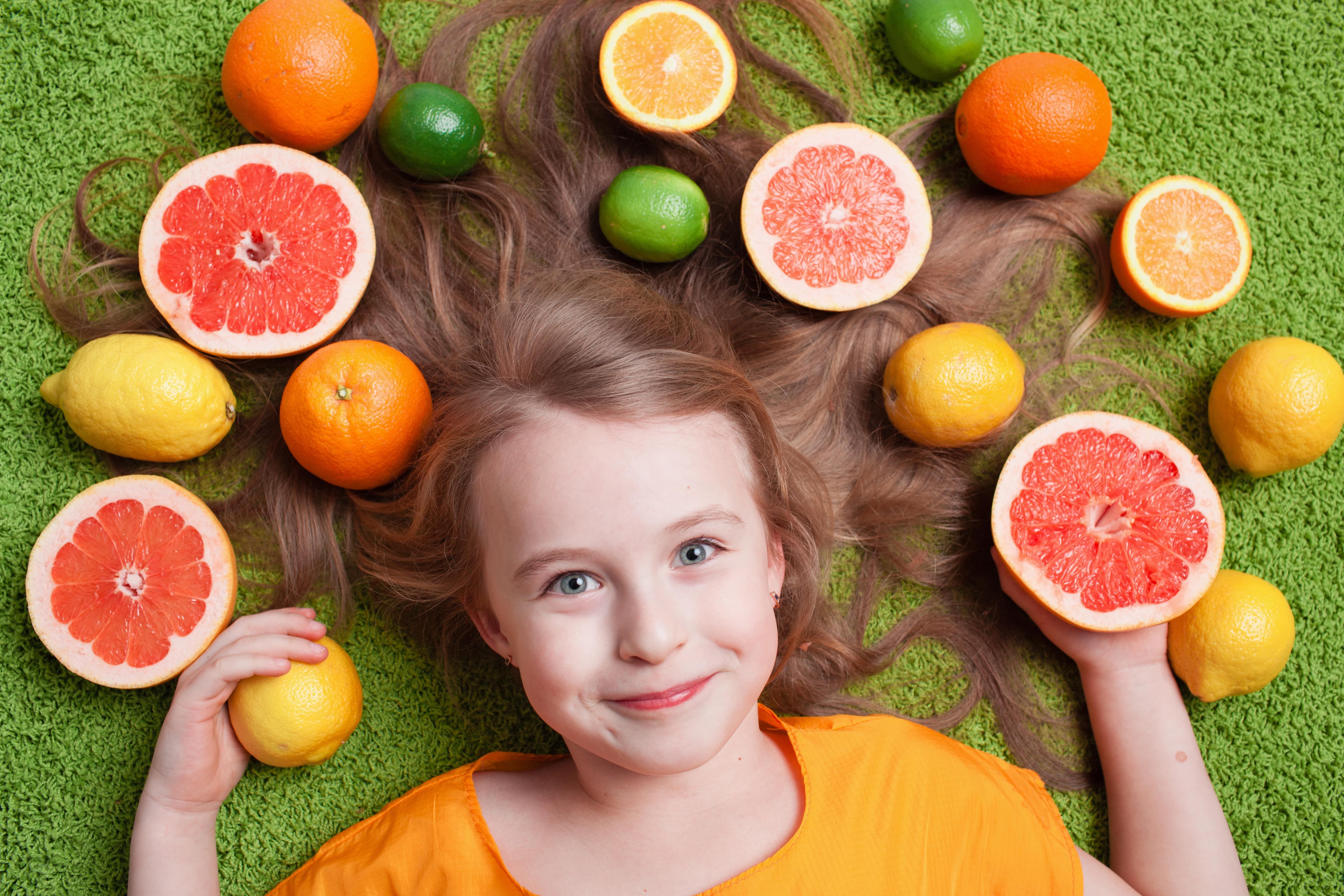 Blonde girl laying down on a green background with her hair spread out and surrounded by citrus fruits