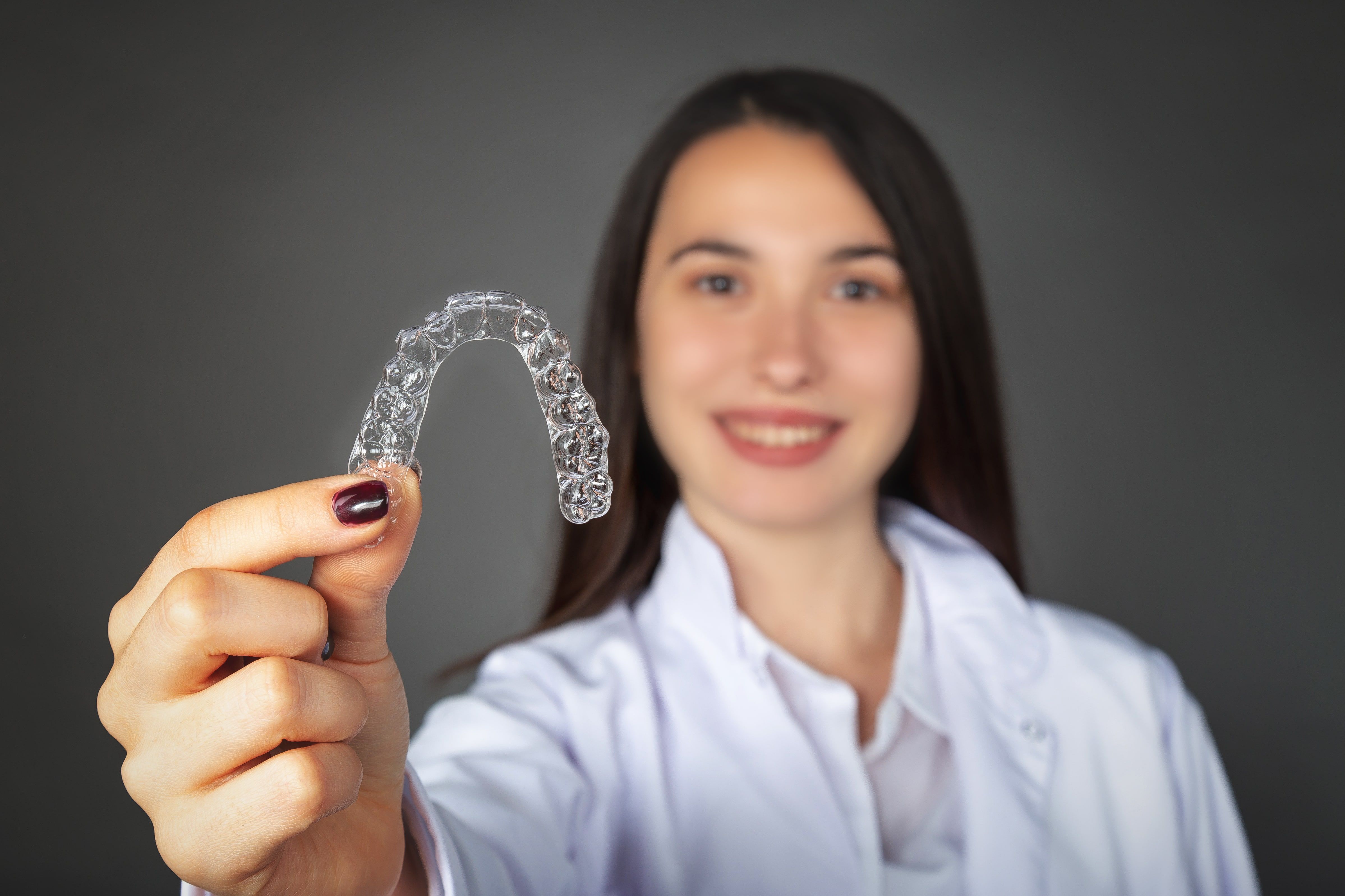 Young woman standing at a distance smiling and holding up an invisalign aligner