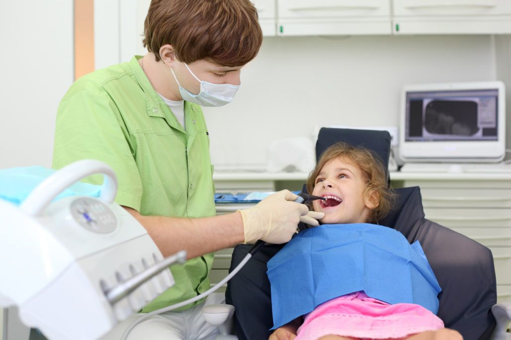 little girl sitting in dental chair smiling at the male hygienist in green scrubs
